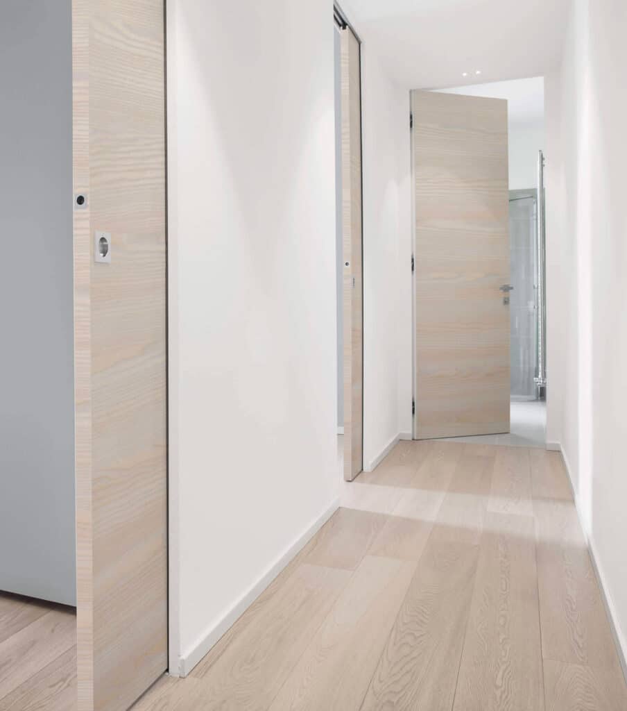 Porte coulissante invisible avec porte essence de bois dans ce couloir moderne et sophistiqué. Une porte battant affleurante complète le look Syntesis
